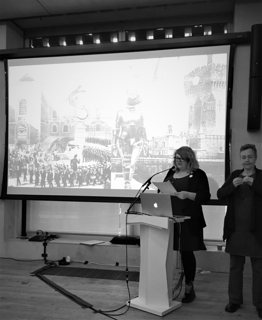 Black and White image of artist Lauren Saunders standing at a podium. The large screen behind her shows two surreal collages she created.